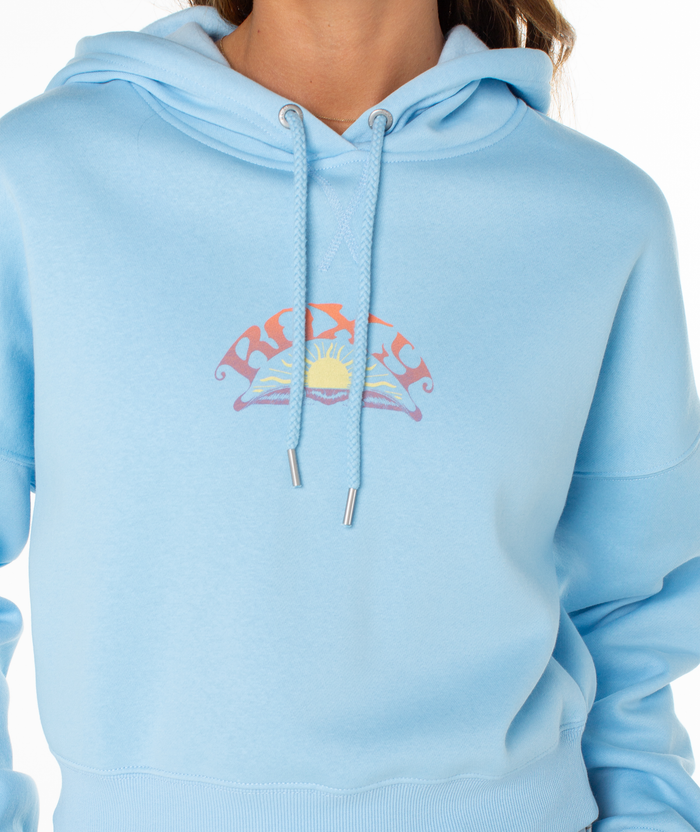 A woman wears the Roxy Breakwater Hoodie in light blue, featuring a colorful sun graphic and logo on the chest. The cropped, relaxed-fit cotton blend hoodie is shown with only her lower face and upper torso visible.