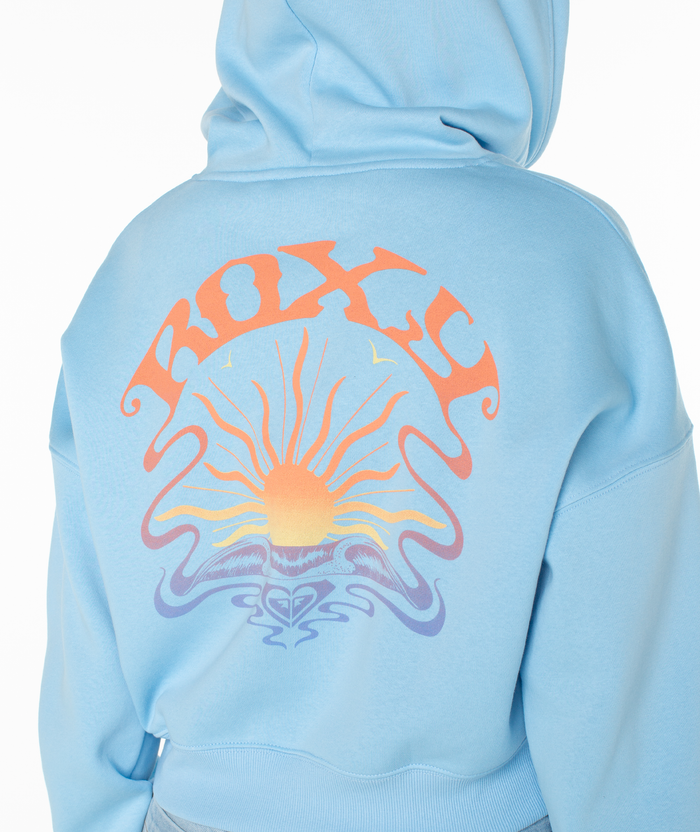Person wearing the ROXY Breakwater Hoodie—a light blue, relaxed cotton-blend hoodie with a cropped fit, featuring bold orange "ROXY" above a colorful sunrise and wave graphic on the back.