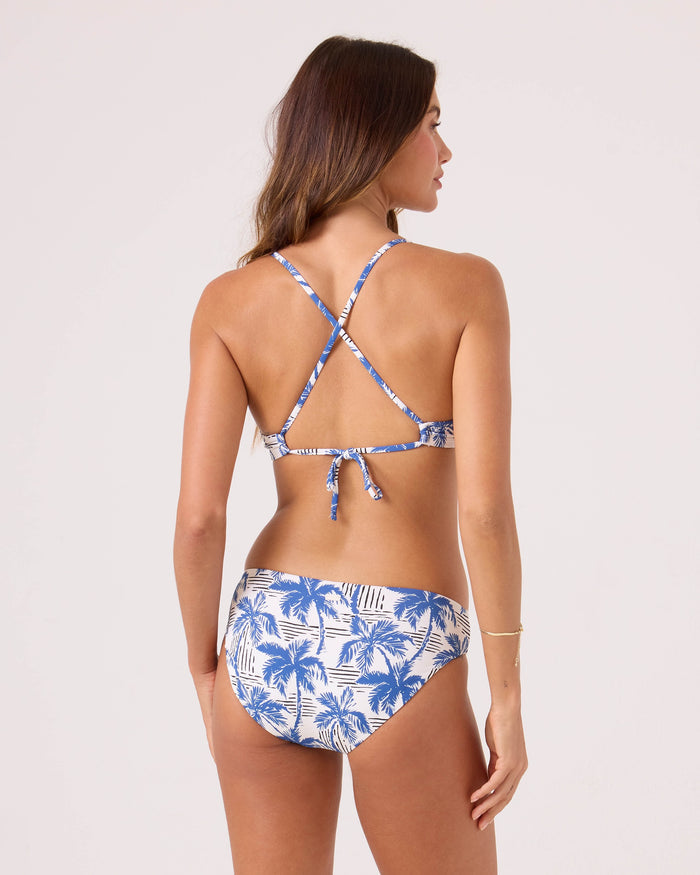 A woman with long brown hair and light skin wears the Roxy Breezy Does It Athletic Triangle Bikini Top, featuring a blue and white palm tree print, crisscross back straps, and a tie-back, standing against a plain background.