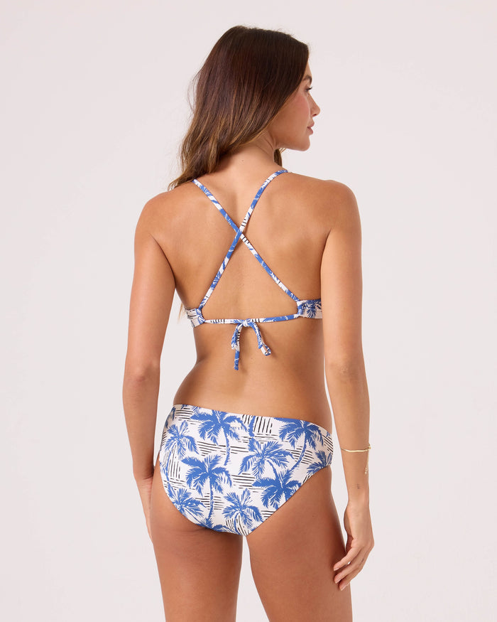 A woman stands with her back to the camera, wearing the Roxy Breezy Does It Hipster Full Bikini Bottom in a blue and white palm tree print, paired with a crisscross-strap top. Confident coverage against a light background—ideal for splash sessions.