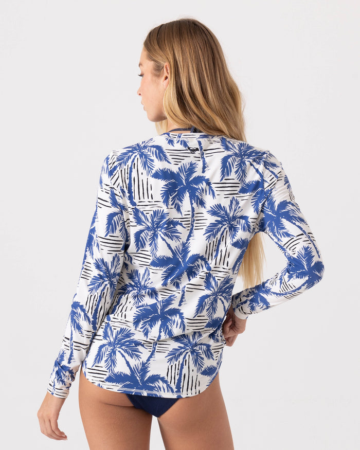 Woman with long blonde hair wears the Roxy Breezy Does It Sea Skippin Rashguard, featuring a blue and white palm tree print, paired with dark bikini bottoms. Shown from the back against a plain background.