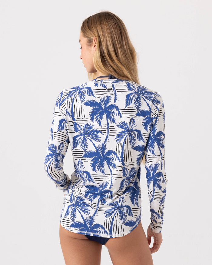 A woman with long blonde hair is seen from behind, wearing the Roxy Breezy Does It Sea Skippin Rashguard with a blue palm tree pattern and dark swimsuit bottoms against a plain, light background.