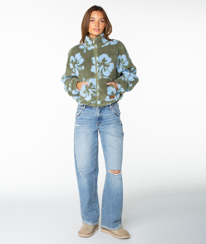 A woman stands facing forward against a plain white background, wearing the Roxy Cold Surf Zip Up—a cropped fit green and blue floral zip-up jacket with a subtle hibiscus print—paired with light blue ripped jeans and tan slip-on shoes.