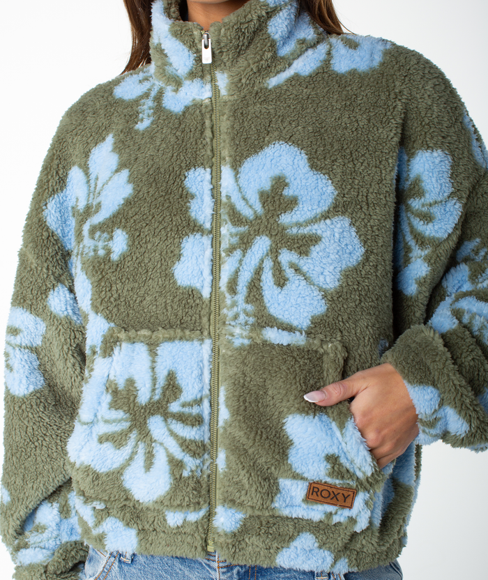 Wearing the Roxy Cold Surf Zip Up, which features a fuzzy olive green and blue floral hibiscus print in a cropped fit, this person has one hand partly in the pocket—displaying the ROXY label—while keeping their face out of view.