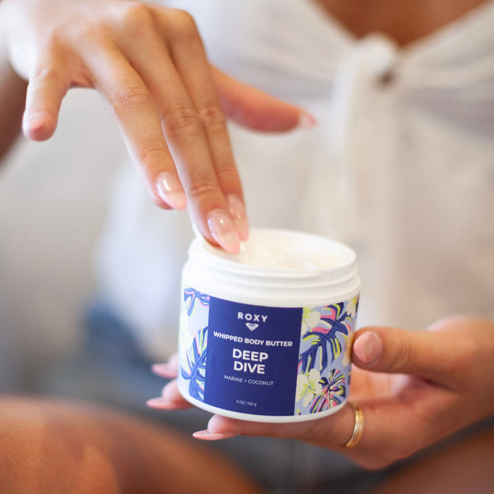 Someone dips their finger into a jar of Roxy Deep Dive Body Butter, featuring a blue and floral label, evoking fresh ocean-air hydration while holding the container in their other hand.