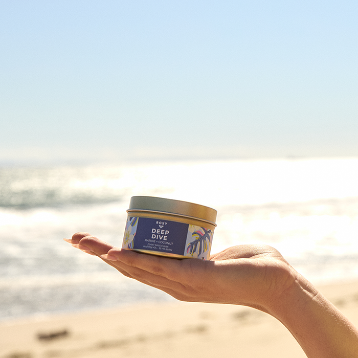 A hand holds a Roxy Deep Dive Palm Tin Candle against a blurred beach backdrop with waves and sun, evoking creamy coconut and refreshing marine breeze scents.