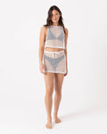 A woman stands barefoot against a white background, wearing Roxy's Don’t Mesh With Me Skirt Cover Up in Egret—a sheer mesh two-piece with a sleeveless crop top and pull-on skirt styled over gray undergarments.