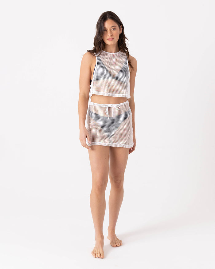 A woman stands barefoot against a white background, wearing Roxy's Don’t Mesh With Me Skirt Cover Up in Egret—a sheer mesh two-piece with a sleeveless crop top and pull-on skirt styled over gray undergarments.