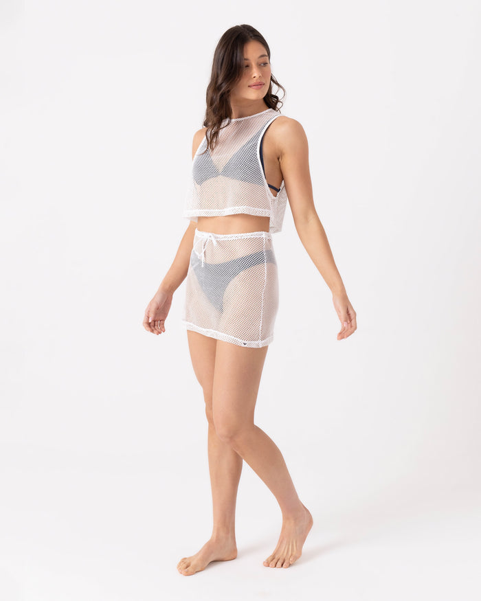 A woman wears the Roxy Don't Mesh With Me Skirt Cover Up—a sheer white mesh sleeveless crop top and matching skirt over a gray bikini—standing barefoot against a white background for a stylish, beach-ready look.