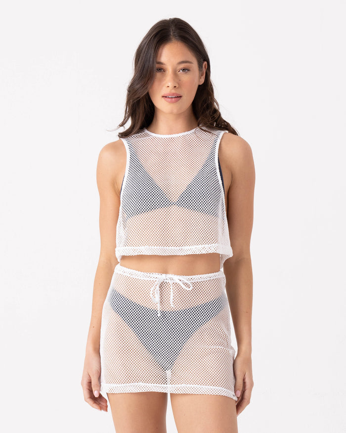 A woman with long, wavy dark hair stands against a white background, wearing Roxy's Don't Mesh With Me Skirt Cover Up and a matching white mesh crop top over a black bikini. She faces forward with a neutral expression, arms at her sides.