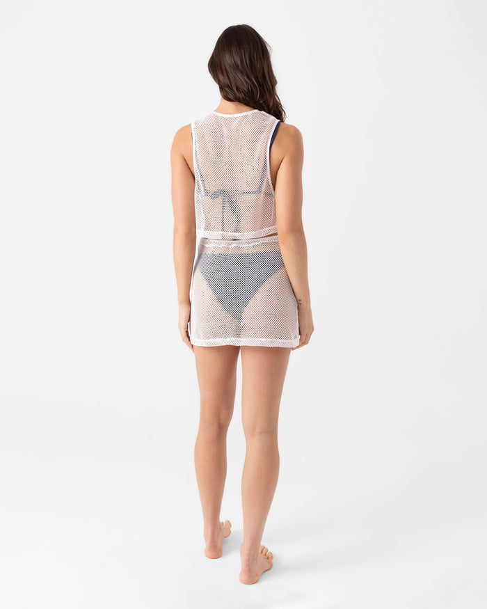 A woman, barefoot and facing away against a white backdrop, models the Roxy Don't Mesh With Me Skirt Cover Up—a sheer white mesh skirt layered over a dark bikini for a beach-ready look.