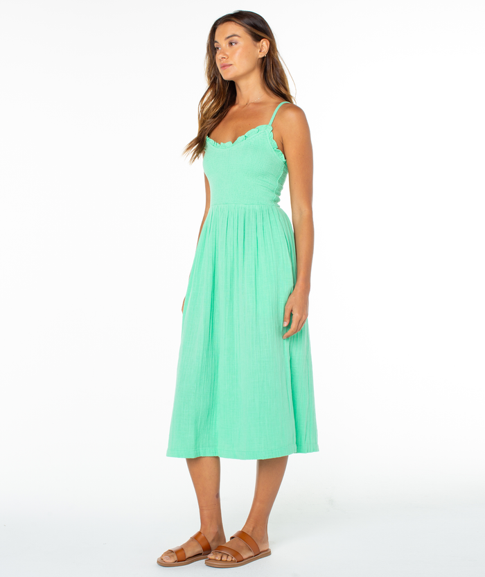 A woman stands against a white background wearing the Roxy Downriver Midi Dress—a light green, coastal style sleeveless V-neck dress with thin straps—paired with tan sandals. She faces slightly to the side, arms relaxed.