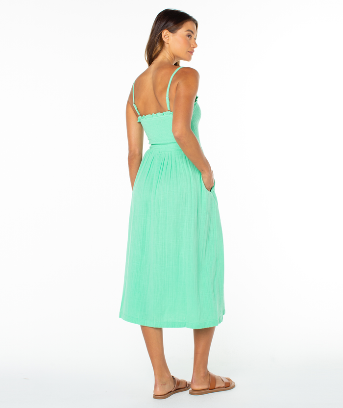 A woman stands facing away in the Roxy Downriver Midi Dress, a light green, sleeveless V-neck with thin straps and sandals, capturing relaxed coastal style against a plain white background.