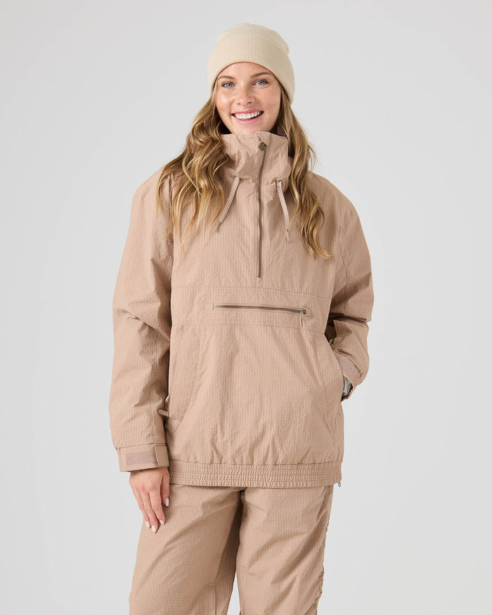 A woman stands against a plain light gray background, smiling with her hand in her pocket, wearing the Roxy Downtown Anorak in Chanerelle—a light brown pullover snow jacket crafted from recycled nylon with a front zipper pocket—paired with matching pants and a beige beanie.