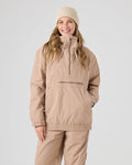 A woman stands against a plain light gray background, smiling with her hand in her pocket, wearing the Roxy Downtown Anorak in Chanerelle—a light brown pullover snow jacket crafted from recycled nylon with a front zipper pocket—paired with matching pants and a beige beanie.