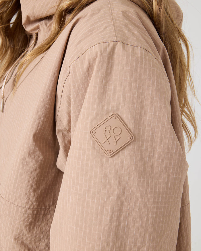 A close-up of a person wearing the ROXY Downtown Anorak, a light brown textured women’s snowboard jacket featuring a diamond-shaped ROXY patch on the sleeve. Only the upper body and part of the hair are visible, showcasing the quality recycled nylon material.