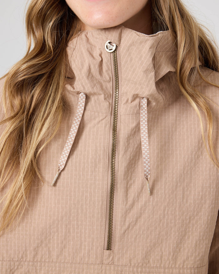 A close-up of a person wearing the Roxy Downtown Anorak, a light brown snowboard jacket with a hood, front zipper, and drawstrings. Crafted from recycled nylon for style and sustainability, the women’s snow jacket partially reveals the wearer’s long, wavy hair.