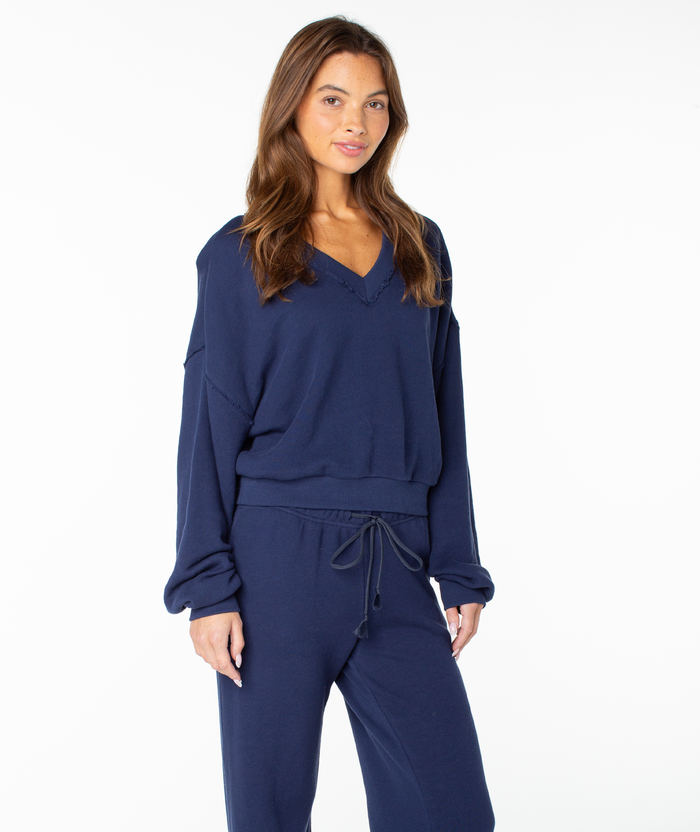 A woman with long brown hair wears the Roxy Dream Thing Top, a navy blue V-neck sweater with a cropped fit, paired with matching drawstring pants. She stands against a plain white background and smiles softly.