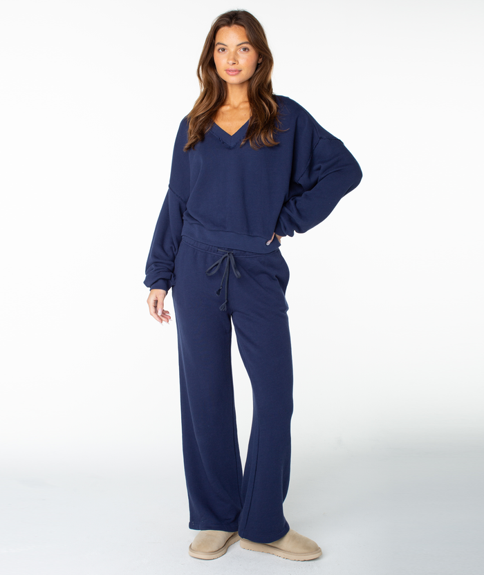 A woman with long brown hair stands facing forward against a plain white background, wearing the Roxy Dream Thing Top—a loose-fitting, navy blue V-neck loungewear top—paired with matching drawstring pants and beige slippers.