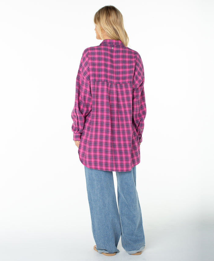 A person stands facing away, wearing the Driftwood Top by Roxy—a cozy cotton-blend shirt in pink and purple tropical plaid with long sleeves—paired with light blue wide-leg jeans, set against a plain white background.