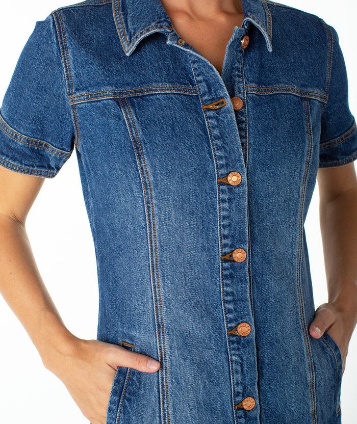 A person wears the Roxy Dusk After Dark Dress, a short-sleeved, dark wash denim style with copper buttons and side pockets. The cropped image focuses on the torso and upper arms, showcasing this fall essential.