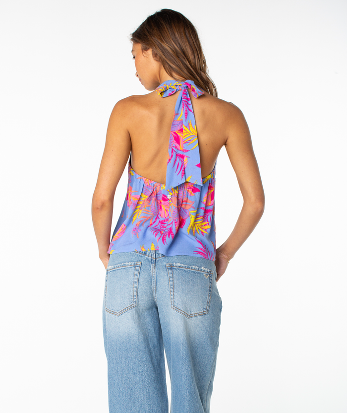 A woman is seen from the back in a colorful floral Roxy Fairweather Top, a sleeveless halter design with a deep V neckline that ties at the neck, paired with light blue jeans and set against a plain white background.