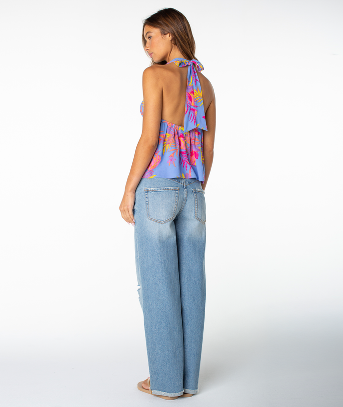 A woman with light brown hair models the Roxy Fairweather Top, featuring a purple floral halter style with a deep V neckline, paired with light blue wide-leg jeans. She is barefoot and photographed against a plain white background.