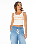 A woman with long brown hair looks to the side, wearing the Roxy Five Palm Stripe Tee—a white baby rib crop top with orange palm trees and text—paired with high-waisted light blue drawstring pants, against a white background in a coastal style look.