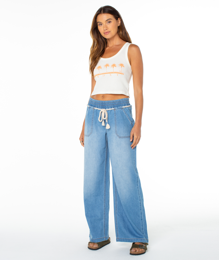 A woman faces forward in Roxy's Five Palm Stripe Tee with an orange palm tree graphic, paired with light blue wide-leg drawstring jeans and dark sandals, showcasing a relaxed coastal vibe against a plain white background.