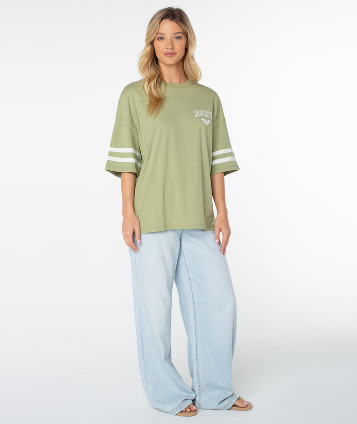 A woman with long blonde hair models the Roxy Game Day Tee—an oversized, boxy-fit light green top featuring retro white sleeve stripes—paired with light blue wide-leg jeans, standing in front of a plain white background.