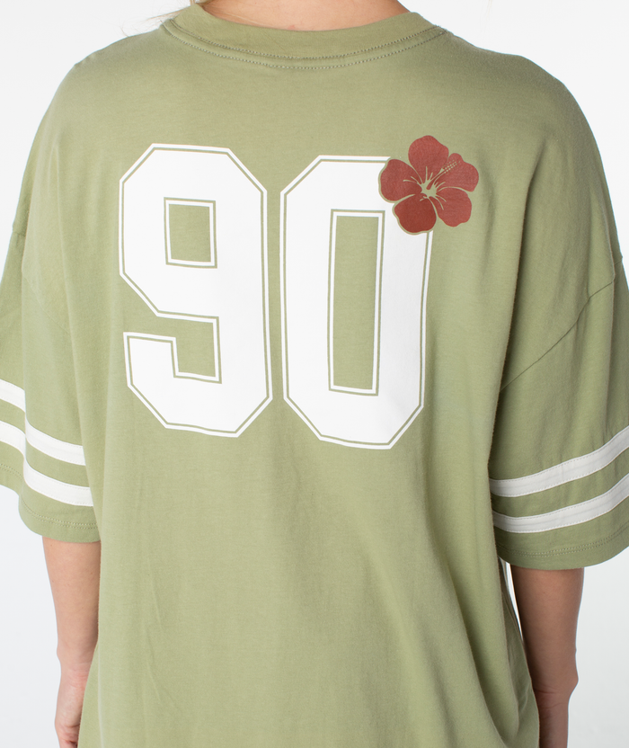 A person wearing the Roxy Game Day Tee in light green, featuring retro-inspired white sleeve stripes, a bold white 90 on the back accented with a red flower graphic, and finished in a boxy fit silhouette that brings cool throwback vibes to any outfit.