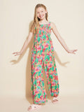 A smiling young girl with long blonde hair wears the Roxy Girls' Coastal Breeze Jumpsuit, which features a smocked bodice. She pairs it with light pink sandals and stands against a plain beige background with her arms slightly raised.