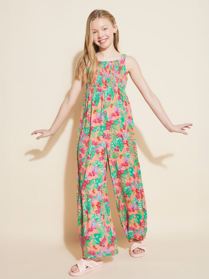 A smiling young girl with long blonde hair wears the Roxy Girls' Coastal Breeze Jumpsuit, which features a smocked bodice. She pairs it with light pink sandals and stands against a plain beige background with her arms slightly raised.