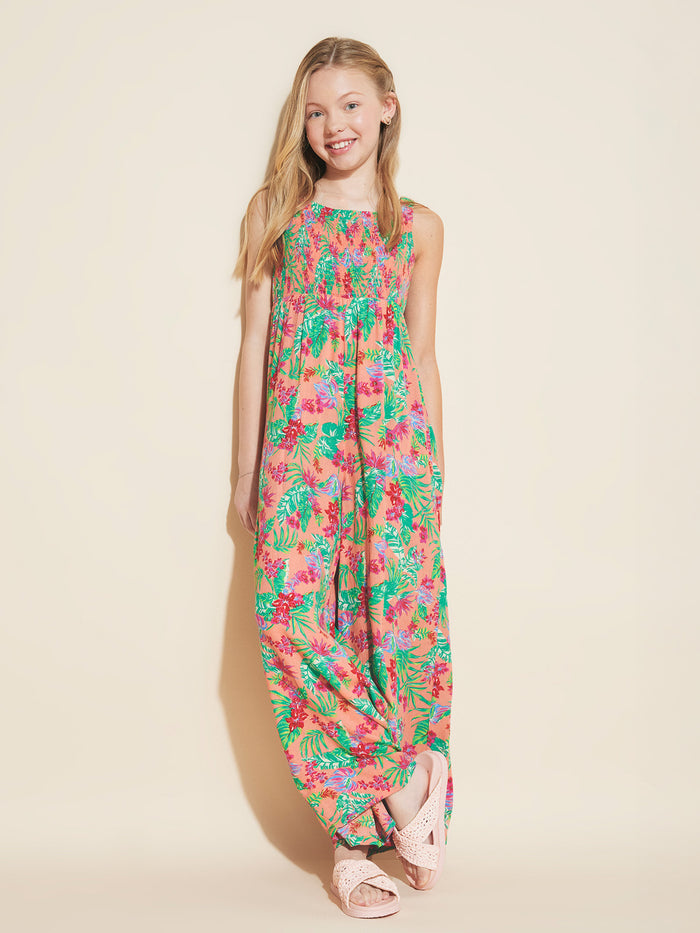 A smiling girl with long blonde hair wears the Roxy Girls' Coastal Breeze Jumpsuit with a smocked bodice and pink sandals, standing against a plain light background with one foot crossed in front of the other.