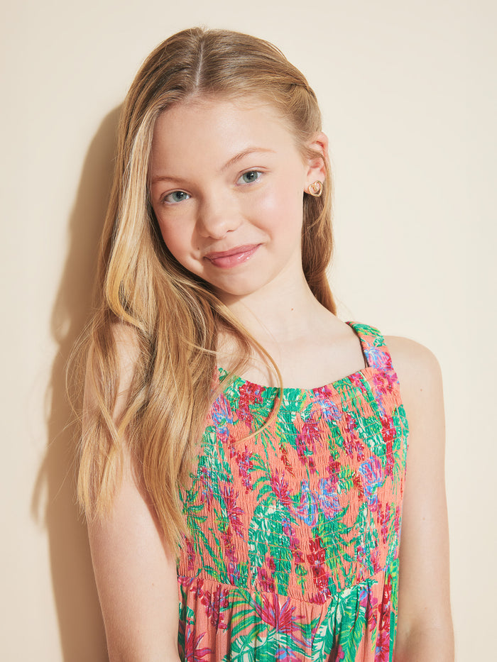 A young girl with long blonde hair wears the Roxy Girls' Coastal Breeze Jumpsuit, featuring a smocked bodice and floral print, as she smiles softly against a light beige background.