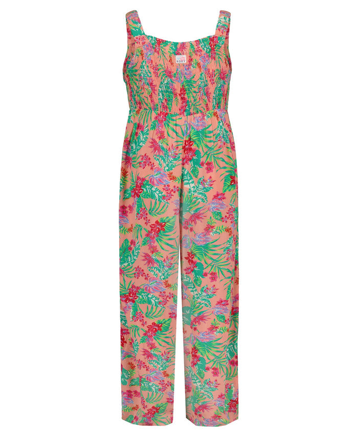 The Girls' Coastal Breeze Jumpsuit by Roxy is a sleeveless viscose jumpsuit with a smocked bodice, wide legs, and a vibrant floral and leafy print in pink, green, red, and purple on a light pink background.