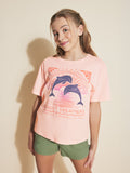 A smiling girl with light brown pigtails wears a Roxy Girls' Ocean Life Oversized Tee with a dolphin graphic and Sunset Dreamers text, paired with green shorts, standing against a beige background.