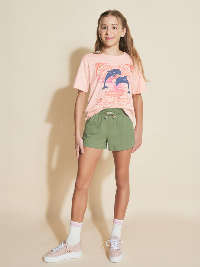 A young girl smiles in a Roxy Girls' Ocean Life Oversized Tee, featuring a dolphin design, green shorts, white socks, and pink sneakers, against a beige background.