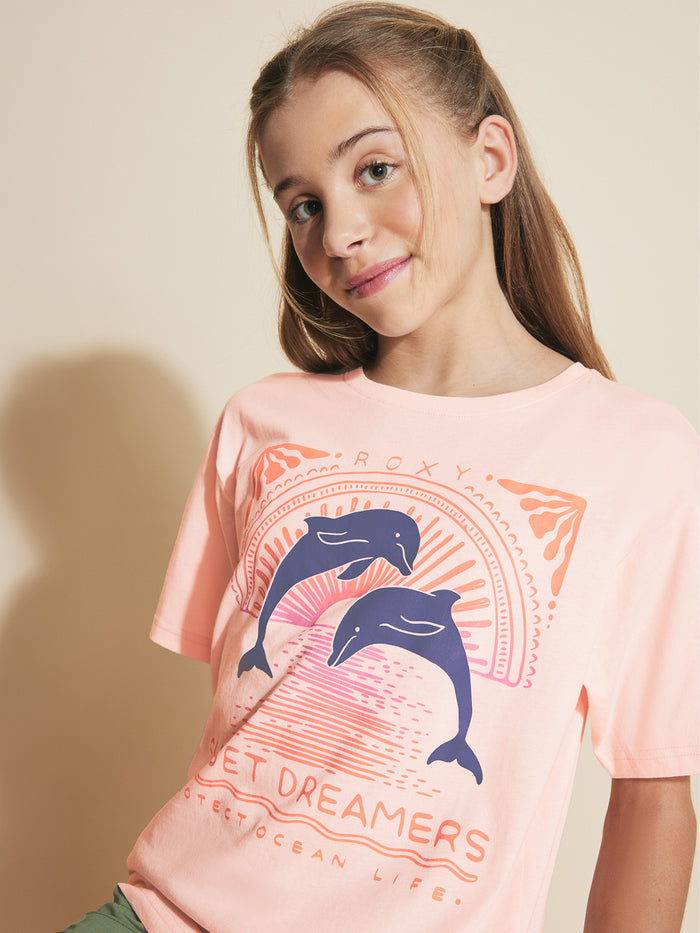 A young girl with long brown hair wears a Roxy Girls' Ocean Life Oversized Tee featuring two dolphins and the words "Secret Dreamers" and "Protect Ocean Life," standing against a beige background and smiling slightly.