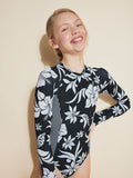 A smiling girl with blond hair pulled back poses against a beige background, wearing the Roxy Girls' Oversized Floral Onesie Swimsuit made from UPF 50+ fabric, standing confidently with one hand on her hip.