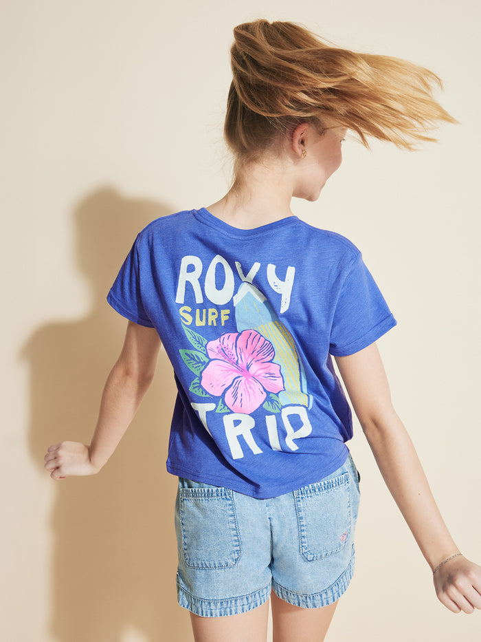 A blonde girl twirls in the ROXY Girls' Surf Trip Tee, made from soft cotton jersey with a pink flower graphic on the back, paired with light denim shorts, set against a beige background.