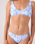 A woman models the Roxy Golden Glow Fixed Lace Hipster Full Bikini Bottom, a light blue recycled stretch bikini with white floral patterns and a scoop neckline, glowing subtly against a plain light-colored background.