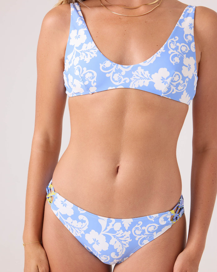 A woman models the Roxy Golden Glow Fixed Lace Hipster Full Bikini Bottom, a light blue recycled stretch bikini with white floral patterns and a scoop neckline, glowing subtly against a plain light-colored background.
