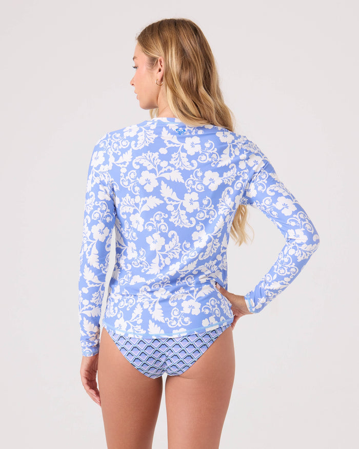 A woman with long, wavy blonde hair is seen from the back, wearing a Roxy Golden Glow Printed Rashguard with blue and white floral prints and matching blue-patterned bikini bottoms made from recycled stretch fabric against a plain background.