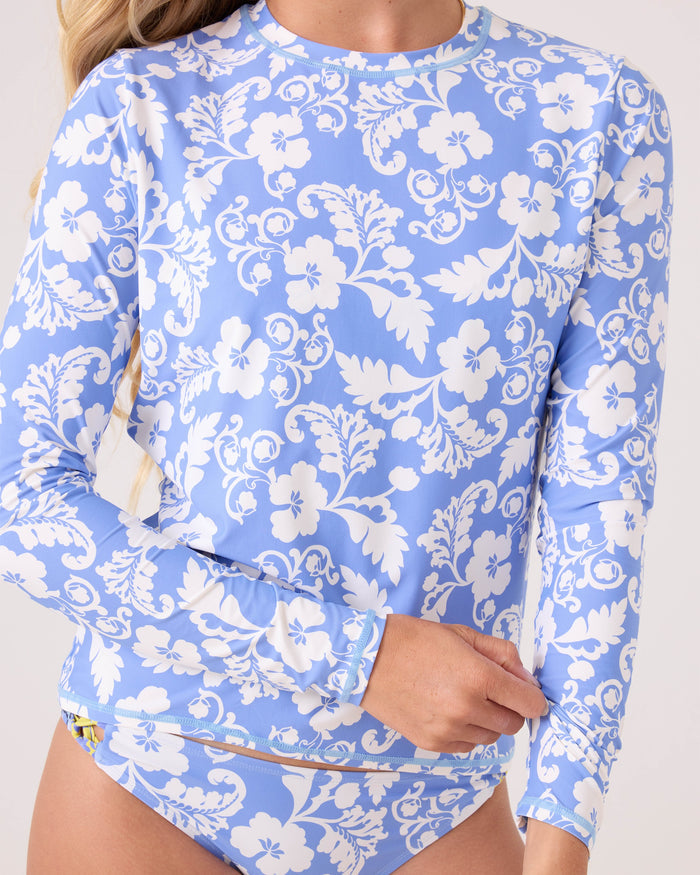 A person wears the Roxy Golden Glow Printed Rashguard, featuring light blue fabric with a white floral pattern. The long-sleeve top is made from recycled stretch material for comfort; their torso and part of their face are visible.