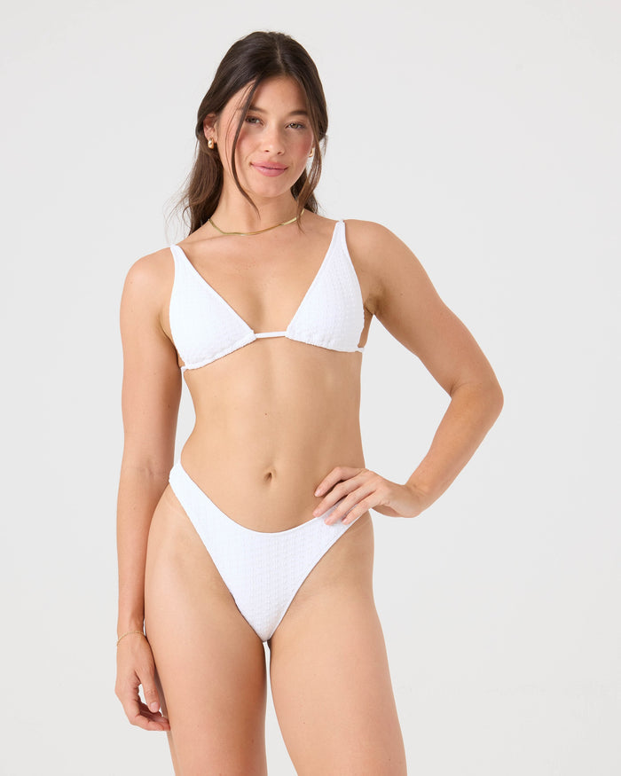 A woman with long brown hair and minimal jewelry stands against a plain light background, smiling slightly as she wears the Roxy Haleiwa Built Up Triangle Bikini Top in white recycled stretch fabric, one hand resting on her hip.