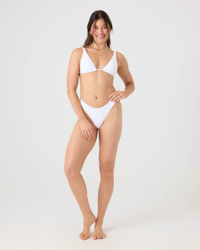 A woman with long brown hair smiles in the Roxy Haleiwa Built Up Triangle Bikini Top, crafted from recycled stretch fabric, standing barefoot against a plain light gray background with her arms relaxed by her sides.