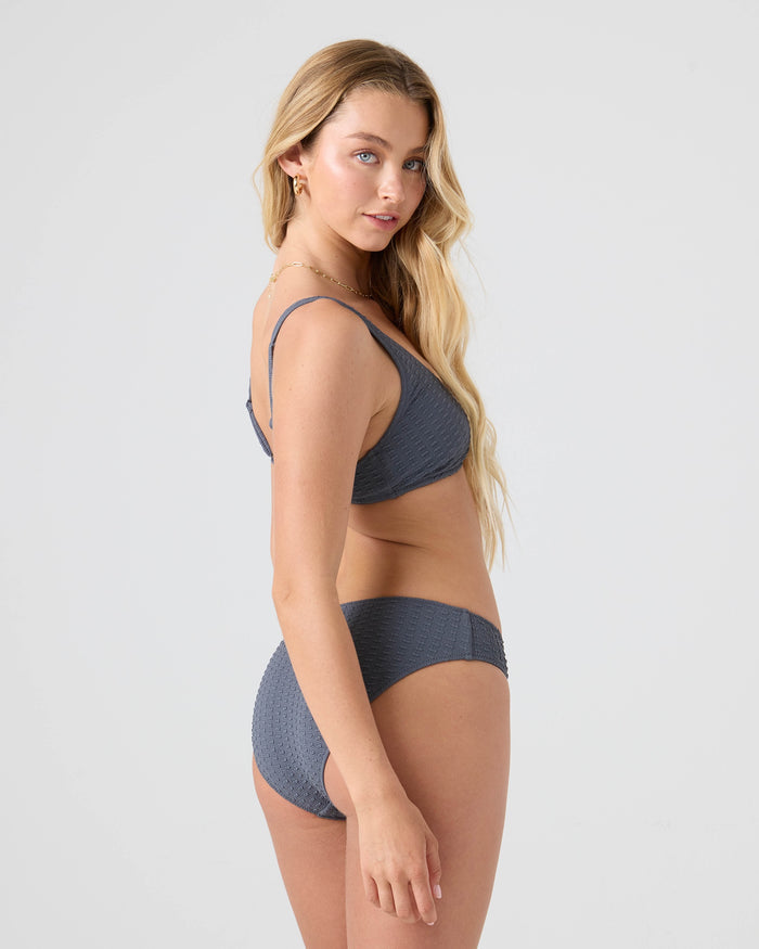 A woman with long blonde hair wears the Roxy Haleiwa Scoop Bralette Bikini Top in textured gray, crafted from recycled stretch fabric, as she stands sideways and looks over her shoulder at the camera against a plain light background.