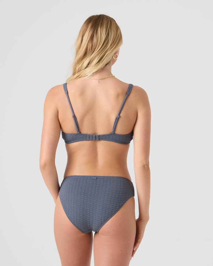 A woman with long blonde hair stands with her back to the camera, arms relaxed at her sides, wearing the textured gray Haleiwa Scoop Bralette Bikini Top by Roxy, made from recycled stretch fabric, against a plain light background.