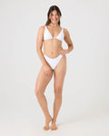 A woman with long brown hair stands barefoot, smiling, and wearing the Roxy Haleiwa Scoop Cheeky Bikini Bottom in white against a plain light gray background.